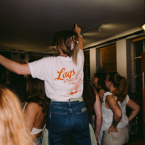 A person wearing a white t-shirt that says The Lags on the back of it in orange. They are daning with their arms in the air. 