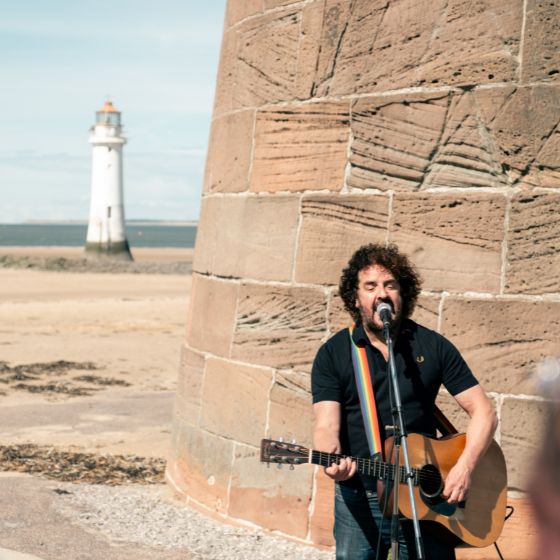 A person playing guitar on a beach with a lighthouse behind them.