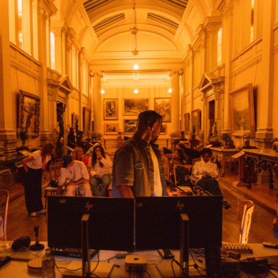 A person stood at two computer screens, turning away from the camera. There is a group of people sitting watching them. The room they are in is inside a museum.