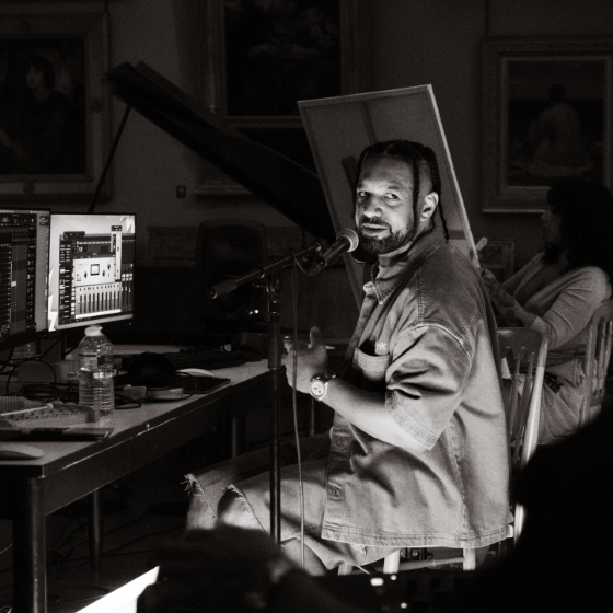 A person sitting at a computer screen looking towards the camera with a microphone.