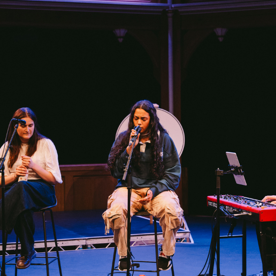 A person sitting on a stool on a stage singing into a microphone. 