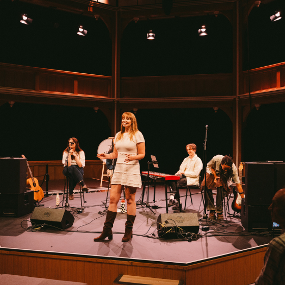 A person stood on a stage in a theatre with people sitting behind them with instruments. 